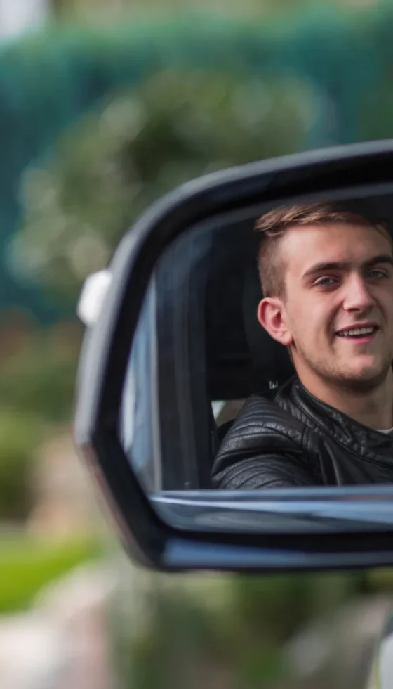 Young teen's reflection in driver's side mirror of his car wearing a seat belt and smiling