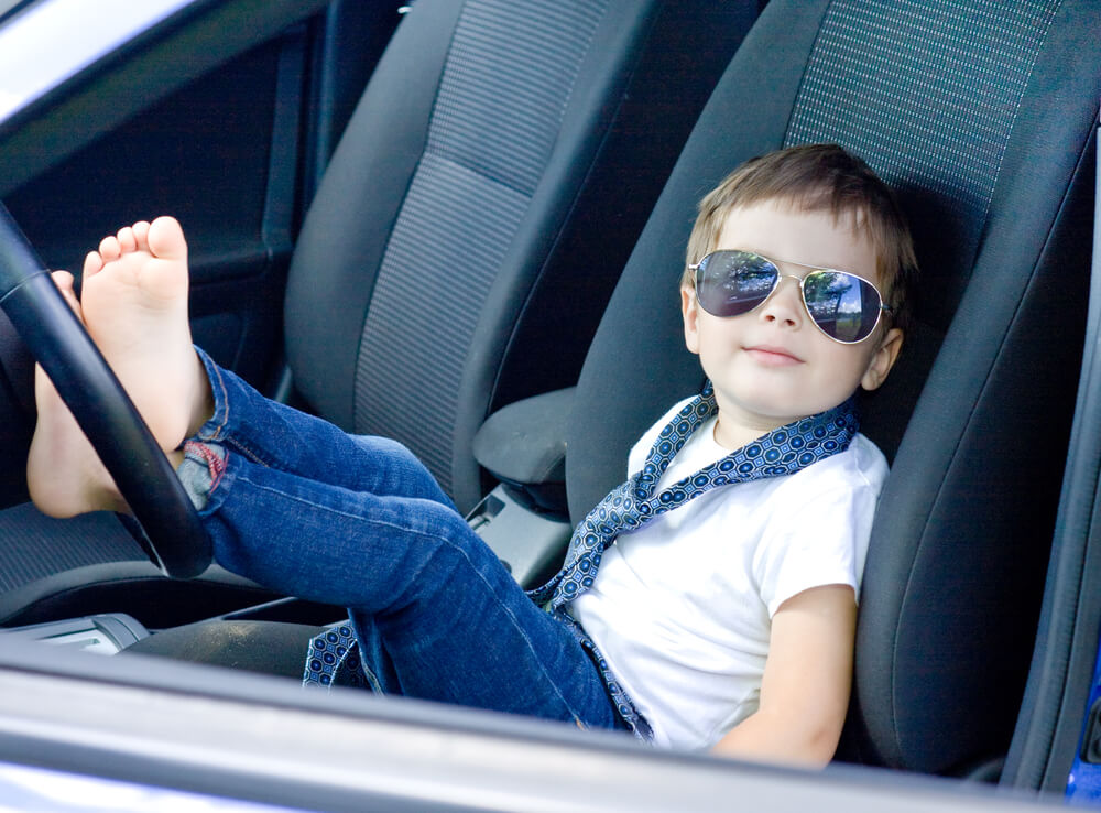 Little boy in sunglasses sitting in a car with his bare feet on the steering wheel wearing a tie - cheap car insurance.