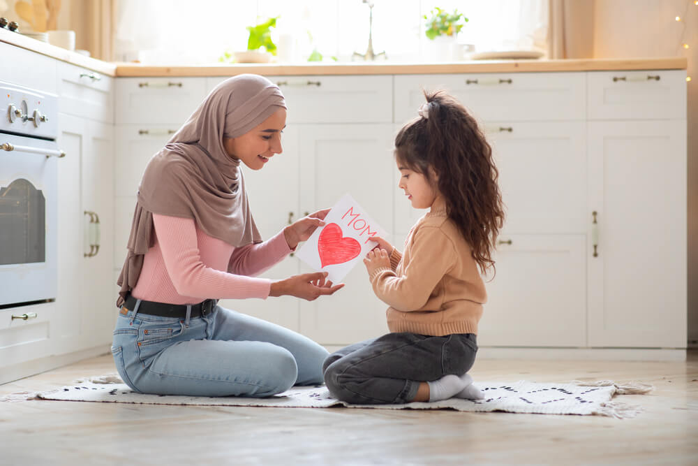 Happy Mother's Day. Little girl greeting her Muslim mom in hijab with gift card with big red heart at home, surprised Islamic mommy and cute daughter sitting together on floor in kitchen, copy space.