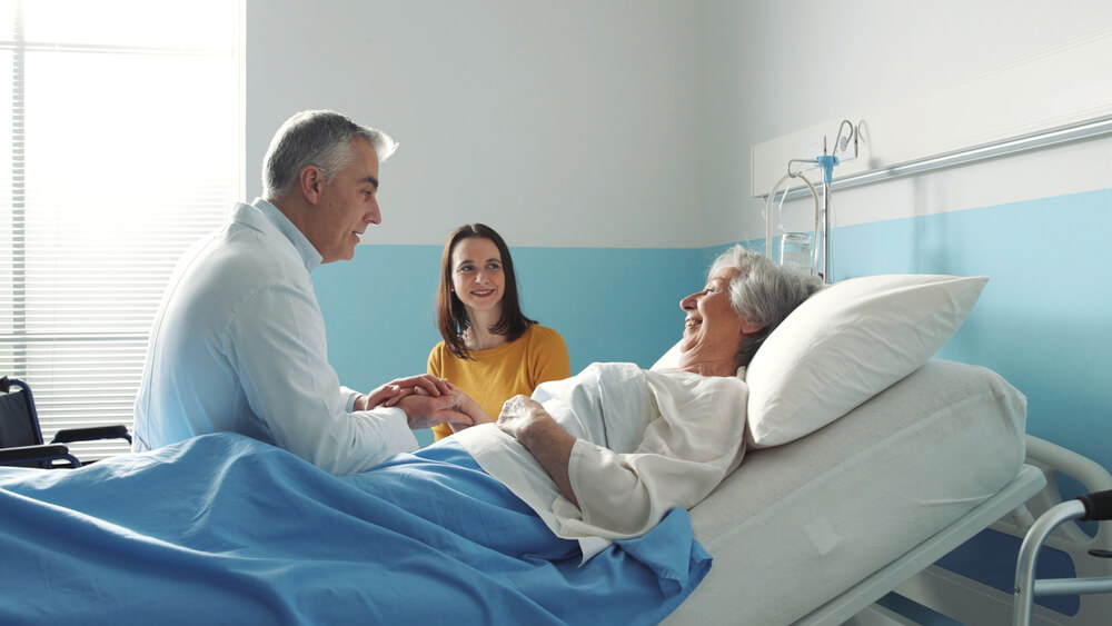 An older woman lies in a hospital bed while her doctor sits on the edge of the bed and talks to her and her daughter looks on - health insurance in California.