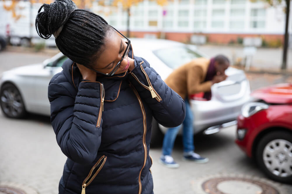 Woman holds her hurt neck while man in the background examines damages after a car accident - cheap car insurance.