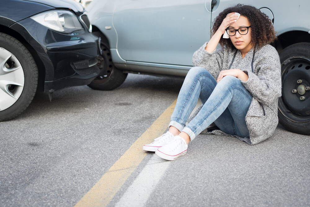 A young woman sits on the ground near her car after a car accident with a cloth to her forehead.