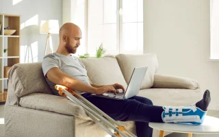 Man with injured leg after a car wreck sits on sofa at home work online on computer.