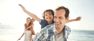 family enjoying the sun on the beach and son with arms up acting like he’s flying on dad’s back