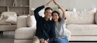 Happy Hispanic 35s couple joining hands under heads showing roof symbol sit on floor in living room