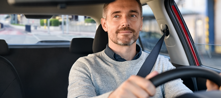 Mid adult man smiling while driving car and looking at mirror for reverse. Happy man feeling comfortable sitting on driver seat in his new car. Smiling mature businessman with seat belt on driving.